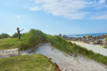 Obraz premium Interested boy taking a picture at a beach at a sunny and windy summer day, standing on a dune