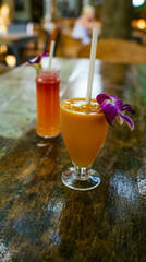 A healthy orange smoothie with a red smoothie in the background in a tropical cafe. The cool fresh juices or cocktails with straws and decorative flowers on the table.