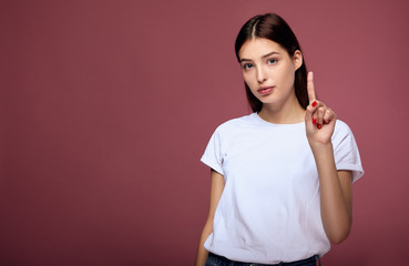 Cheerful young lady raises finger above stands against pink background.