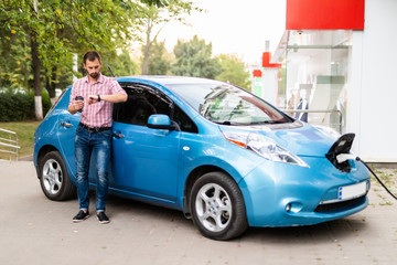 Young handsome man look at watch near his electric car charging at station