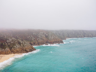 Wide aerial view of the rocky granite cliffs along the foggy coasts of Porthcurno Beach. Cornwall, United Kingdom. Travel and nature. - 310677264