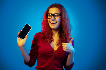 Caucasian woman's portrait isolated on blue studio background in neon light. Beautiful female model. Concept of human emotions, facial expression, sales, ad. Showing blank phone screen. Thumb up.