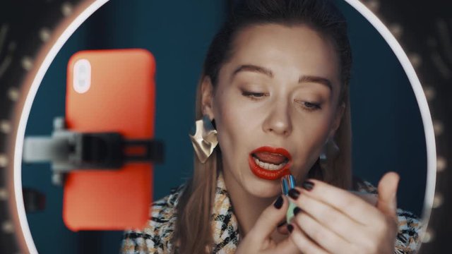 Attractive woman fashion blogger sits in front of ring light and orange smartphone, applying red lipstick, showing technique of makeup, recording tutorial video. Shot on 4K RED camera.