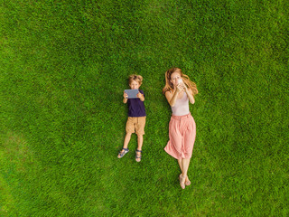 Obraz premium Mom and son are lying on the grass in the park. Mom looks at the phone, son looks at the tablet. Photos from the drone, quadracopter