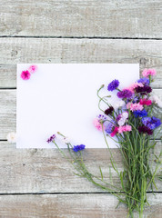 Colorful bouquet of summer garden flowers. Cornflowers with blank paper for greeting message on old shabby wooden table. Vintage floral background. Floral mock up with multicolored flowers. Copy space