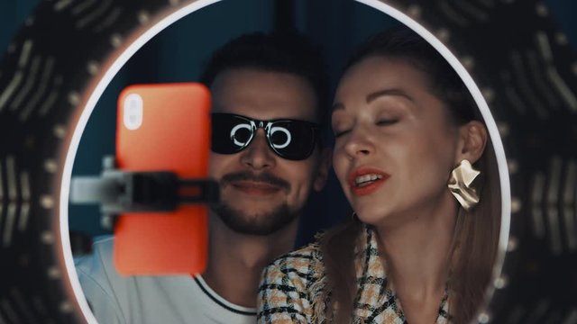 Good-looking man in sunglasses and blonde woman fashion blogger are talking with smile in front of ring light, looking at camera of smartphone, shooting video for their channel. Shot on 4K RED camera