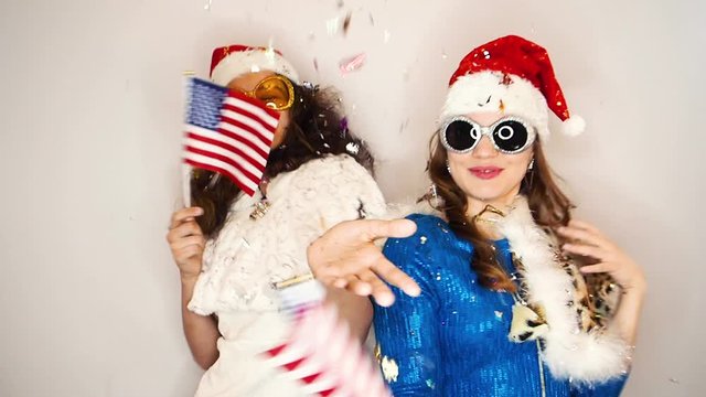 Two dancing girlfriends in a Santa Claus hats waving the flags of USA in the rain of candy on a light background. New Year holidays concept in the world. Slow motion