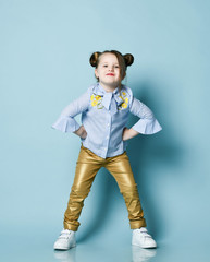 Playful frolic kid girl in blue shirt and gold leather pants is cool posing with her hands on her...