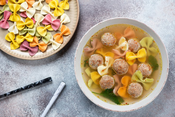Soup with meatballs and pasta over beige stone background with raw farfalle, above view, studio shot