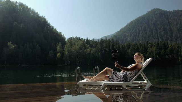 Woman shooting on handheld film gimbal stabilization for smartphone. Girl rest, relax, lie sunbed on pier in sunglasses, make selfie. Lady blogger broadcast video blogging vlogging. Take photo video.
