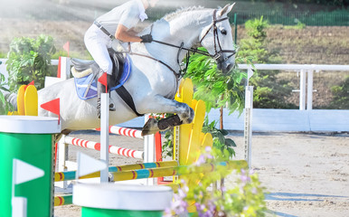Horse and rider in light uniform performing jump at show jumping competition. Beautiful white horse...