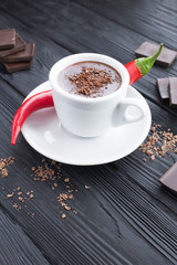 Hot chocolate in the white cup and red peper chili on the black wooden background. Location vertical.