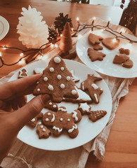 Hand holding gingerbread christmas tree cookie with icing on background of wooden table with lights, candles and holiday decorations. Festive christmas tree cookie