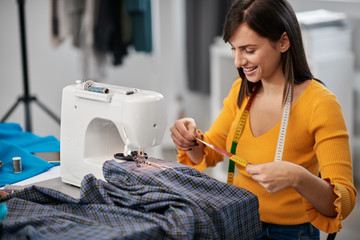 Focused charming caucasian fashion designer sitting in her studio and measuring textile for...