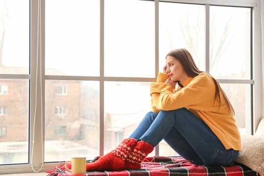 Young Woman In Winter Clothes Sitting On Window Sill At Home