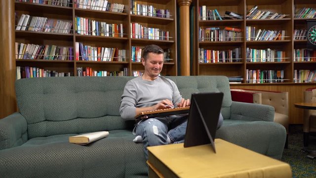 Smiling man types text with retro keyboard and Fire HD tablet on couch in home book library - panning horizontal, wide