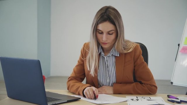 American businesswoman working with laptop ux design and writing sitting at table in office. Young confident woman watching computer screen, write in document at desk in company interior. Female