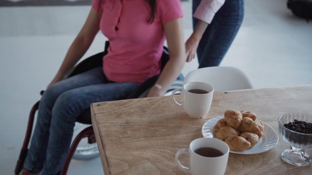Disabled woman sitting in wheelchair and riding with help friend in home room. American paraplegic female moving in medical chair in house interior near table with coffee and food. Concept: recovery
