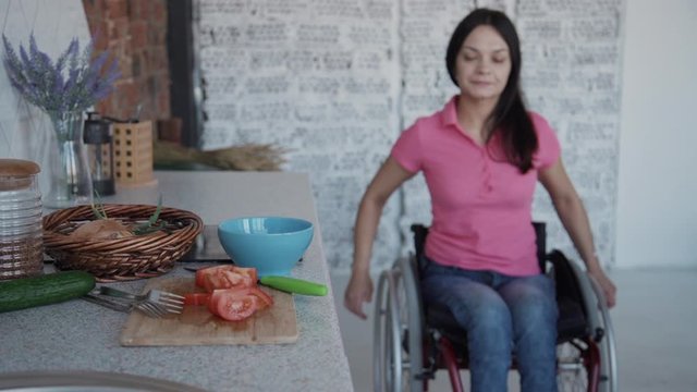 Young paraplegic woman in wheelchair riding to table with vegetable in kitchen room. Disabled female using medical chair moving to desk to cooking vegetarian salad from cucumber and tomato. Lady
