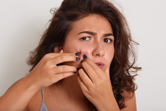 Indoor shot of young woman squeezing pimple on her cheek isoalted over white background, looking at camera with scared facial expression, beautiful girl wearing casual clothes, having dark wavy hair.