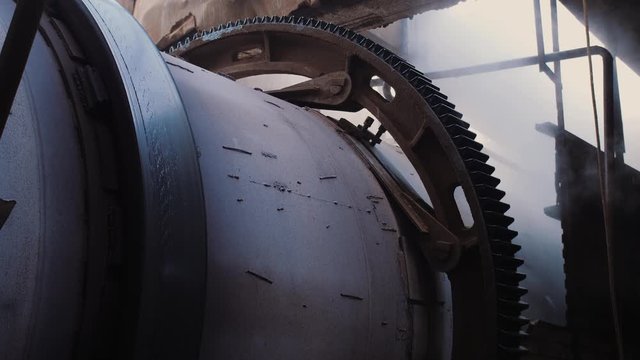 Rotating gear of huge industrial machine, steam on the background. Rotary kiln on a cement plant. Industrial production process of cement clinker or expanded clay. Huge industrial oven on a factory.