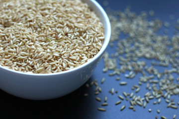 Brown rice grains, uncooked, in a white bowl on a dark surface