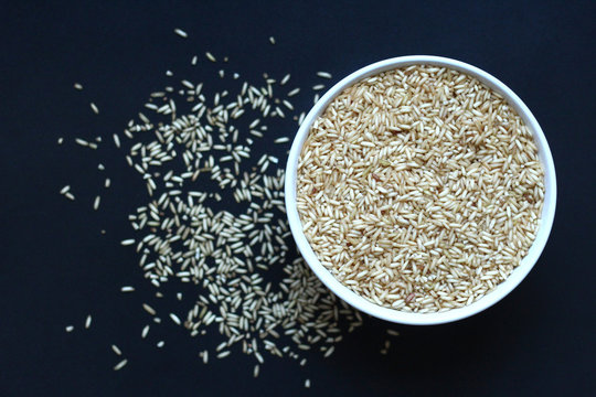 Brown Rice Grains In A White Bowl On A Dark Background, Seen From Above