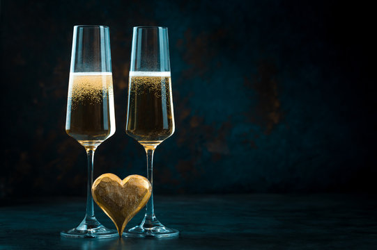 Two elegant romantic glasses with sparkling golden champagne with golden heart against a dark blue background. Valentine's day.