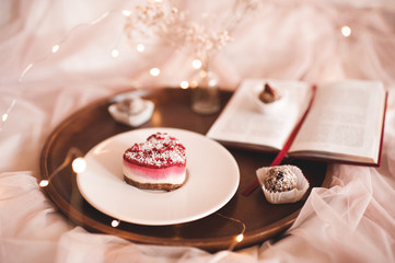 Tasty homemade cake with raspberry on white plate with open book on wooden tray in bed closeup. Good morning. Breakfast.