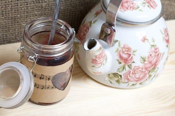 Glass jar of jam wrapped in paper with notes. Heart cut out in paper. Near a white kettle with roses. Stand on the boards, on the background of linen.