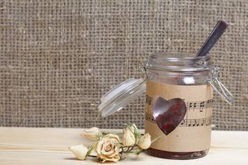 Glass jar of jam wrapped in paper with notes. Heart cut out in paper. Nearby dried roses. It stands on the boards, on the background of linen fabric.
