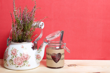 Glass jar of jam wrapped in paper with notes. Heart cut out in paper. Near the kettle with a bunch of heather. It stands on the boards, on a coral background.