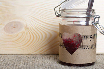 Glass jar of jam wrapped in paper with notes. Heart cut out in paper. It stands on the boards, on the background of linen fabric.