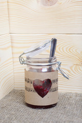 Glass jar of jam wrapped in paper with notes. Heart cut out in paper. It stands on the boards, on the background of linen fabric.
