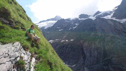 Very steep mountain meadow with emerald green grass exhausted hiker climber man boy is sitting on the edge enjoying the view and relaxing in the Swiss Alps Mountains and glaciers background