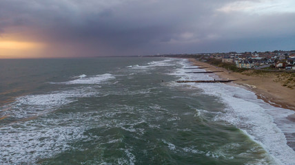 An aerial view of a choppy sea with crashing waves, groynes (breakwater), sandy beach and city in the background under a stormy cloudy grey sky at sunset