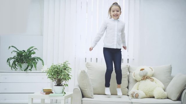 Cheerful little girl is bouncing on the sofa joyfully, smiling. Soft teddy near her.
