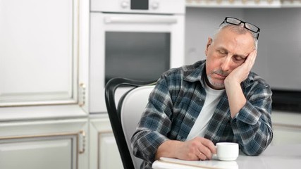 Tired sluggish aging male dozing during drinking tea at white kitchen medium shot