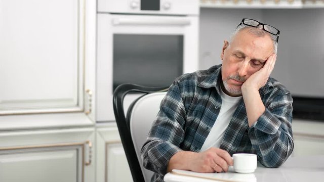 Tired sluggish aging male dozing during drinking tea at white kitchen medium shot