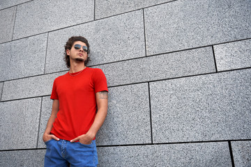 Freestyle. Stylish guy in red t-shirt sunglasses standing isolated on grey posing looking aside cool