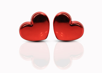Two red plastic hearts lie on the table reflected in the glossy surface on a white background....