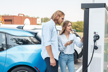 Traveling. Young couple traveling by electric car stopping at charging station girlfriend with...