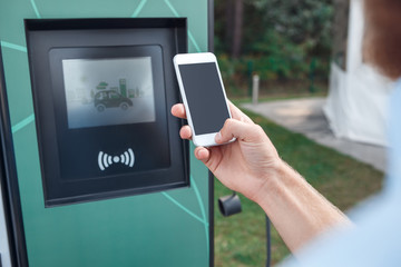 Transportation. Young man traveling by electric car stop at chraging station browsing smartphone close-up copy space paying online looking at machine display