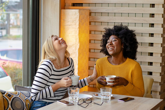 Two multi ethnic girl friends enjoying coffee together in a coffee shop, sitting at a table chatting and laughing loud.