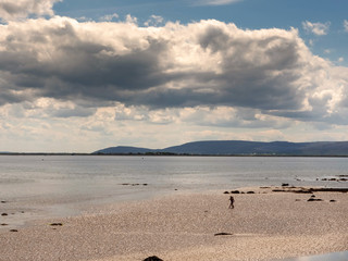 Silhouette of two woman on a sandy beach walking towards water, Galway bay, Burren mountains in the background, Cloudy sky over ocean.