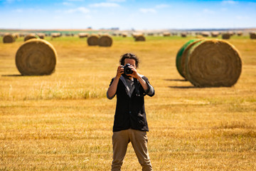 Wide angle view of male photographer standing in an agricultural field with bales of golden hay, shooting at camera with a DSLR camera. 