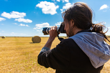 Male photographer with long hair shooting bales of hay in the countryside with professional DSLR camera and expensive looking long lens. 