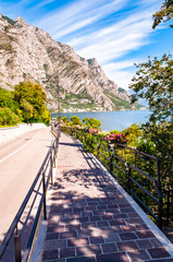 Cozy city street of Limone Sul Garda with paving stone sidewalk, blooming flowers on a metal railings, growing trees with amazing Garda lake and dolomite mountains on the background. Scenic promenade