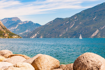 Round boulders lying on the shore of beautiful Garda lake in Lombardy, Northern Italy surrounded by high dolomite mountains. Single classic white sailing yacht floating on the lake
