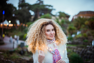 Portrait of smiling young curly blond woman in warm white natural sweater with garland looking at camera with the green park background with bokeh abstract lights. Selective focus. Copy space.